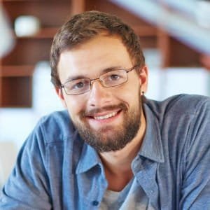 A man with glasses smiling indoors, casual attire, in a professional setting.